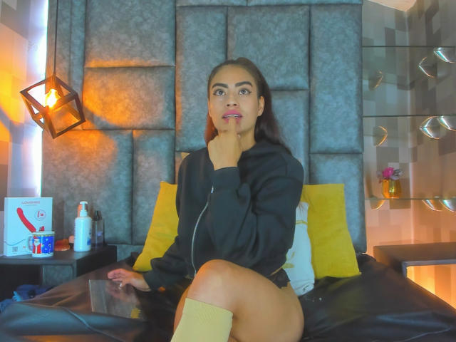 EmilyMitchell with medium brown skin, dark hair, and pink lipstick, wearing a black hoodie, yellow knee socks, and black underwear, sitting on a bed, hand on chin.