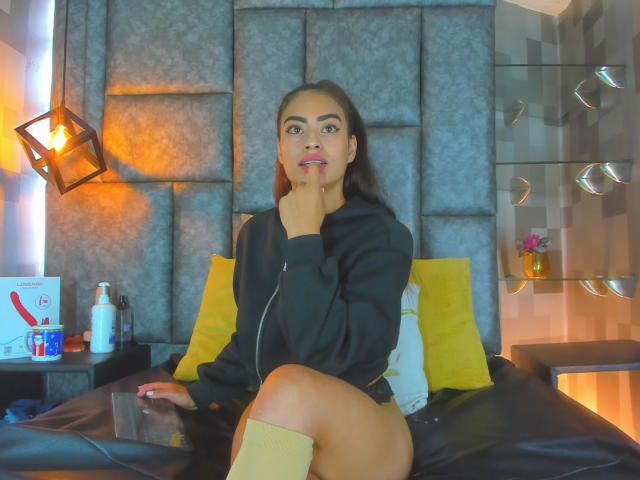 EmilyMitchell with brown hair, black hoodie, yellow socks, and no pants, sitting on a bed, finger on chin, smiling, yellow pillows behind her, bottle and box on side table.