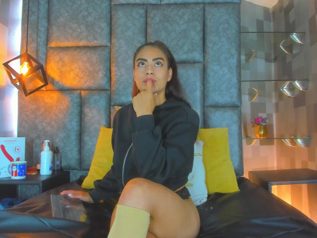 Emily Mitchell, mit dunklem Haar, trägt eine schwarze Kapuzenjacke und gelbe Kniestrümpfe, sitzt auf einem Bett, lutscht an ihrem Finger, wobei ihr Hintern und ihre Vulva kaum sichtbar sind..