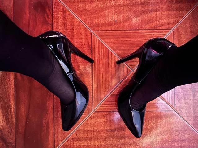 EliseMonet's glanzende zwarte hoge hakken met puntige neuzen en hoge stiletto's, staan op een glanzende houten vloer. Haar benen zitten in zwarte leggings..
