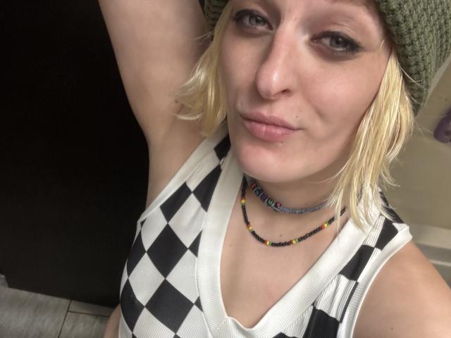 KirryBoby, com cabelo loiro, gorro verde, regata quadricula preta e branca e colar de contas, mostrando a axila, sorrindo, com maquiagem discreta e um pequeno piercing no nariz..