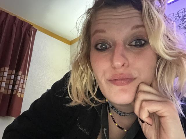 KirryBoby met golvend blond haar, blauwe ogen en een neusring, met een frons en een hand op haar wang, gekleed in een zwart shirt en een ketting, die recht in de camera kijkt..
