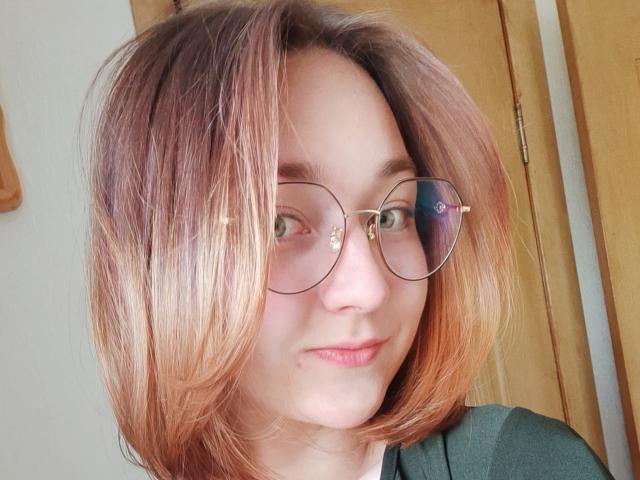 LittleRory mit bräunlich-rosa Haaren, grünen Brillen und einem leichten Lächeln, zeigt ihr süßes Gesicht und ihre grünen Augen. Sie trägt ein grünes Oberteil, im Hintergrund ist eine Holztür zu sehen..