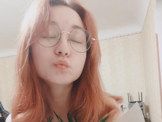 LittleRory, con i capelli rossi, le labbra piene e gli occhiali rotondi, fa spuntare leggermente il suo naso un po' all'insù. Si vede la sua maglietta verde. Luce soffusa, leggermente sfocata..