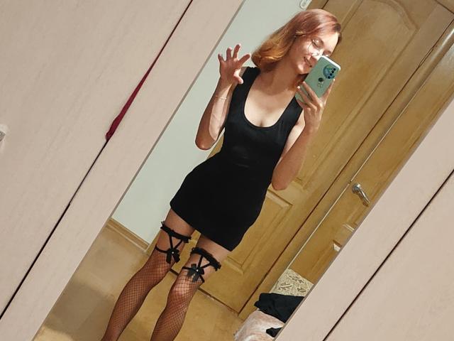 LittleRory, con i capelli rossi, indossa un vestito nero, calze a rete e bretelle nere, sta scattando un selfie allo specchio, mostrando un viso carino e un gesto giocoso con la mano..