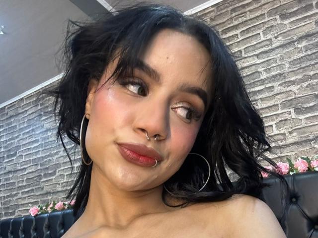 WendyJames has dark, wavy hair, a nose ring, a septum ring, and hoop earrings. She's looking sideways with pink lipstick and flushed cheeks. Her tits are slightly visible.