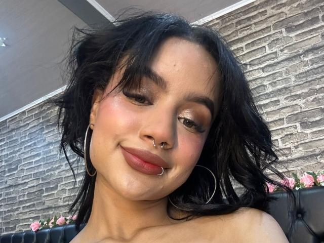 WendyJames has medium brown skin, black wavy hair, and a nose ring, wearing hoop earrings and pink lipstick, with a slight smile, showing her tits and ass.