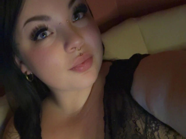MadameMarie-hot with pouty lips, dark lace top, nose ring, and hoop earrings, has her tits slightly visible, and blue eyes looking at the camera, in a dimly lit photo.