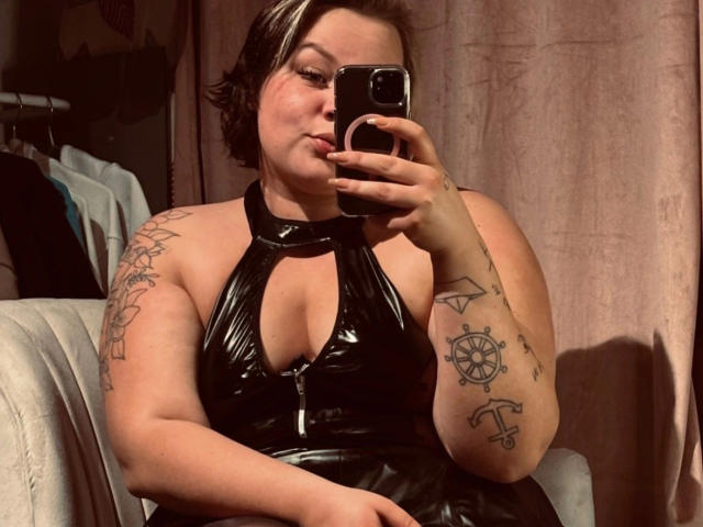 MadameMarie-hot with short black hair, puffy lips, and tattoos, wearing a shiny black halter top, taking a mirror selfie with a phone, showing her big ass.