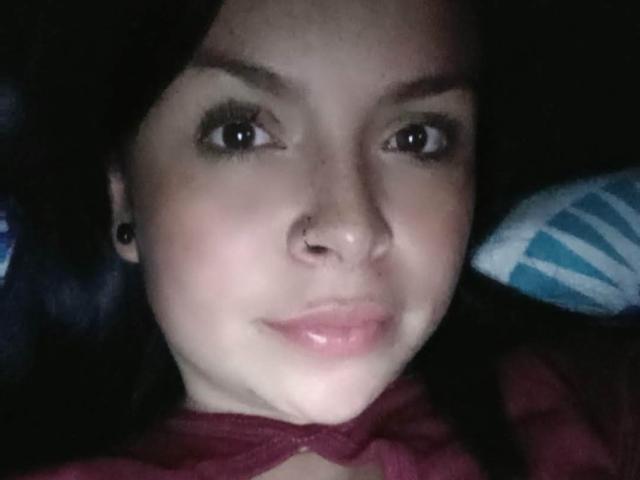 EntoncesIrene with a nose ring, black ear studs, dark hair, and pink glossy lips, looking at the camera with a slight smile in a dimly lit photo.