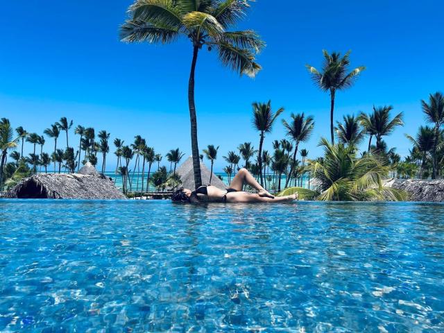 SelenaCarter lies in a clear blue pool, wearing a black bikini. She's sunbathing on her back with legs slightly bent, head turned to the side. Her blonde hair is wet, and she's partially submerged. Tall palm trees with lush green leaves are in the background, and a thatched roof structure is visible to the left. The sky is bright blue. The pool's texture is smooth, with light reflections on the water surface. The overall composition is vibrant and tropical.