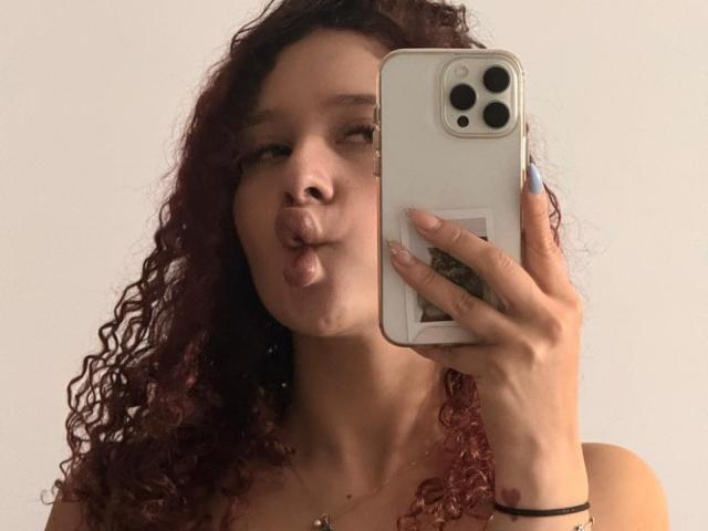 ValeryScot with curly red hair, puffy lips, holding a phone, wearing a necklace, and blue nails, taking a mirror selfie. Her tits are slightly visible.