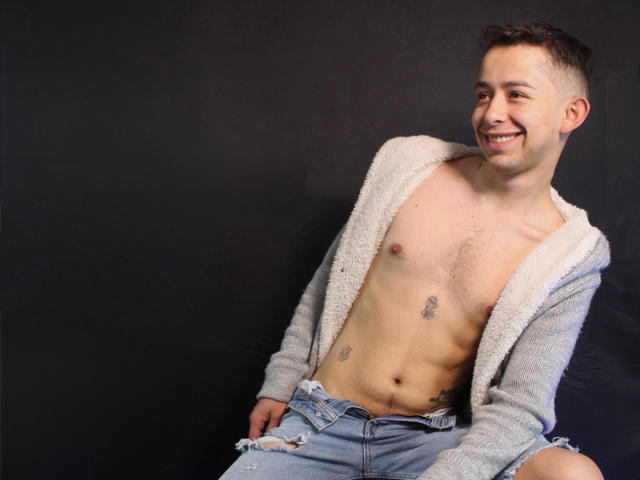 AlecAndrews, shirtless with a gray fuzzy jacket, wearing ripped jeans, smiling, has tattoos on his chest and lower belly, short brown hair, and a slim build.