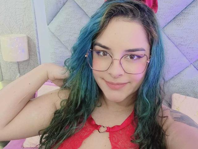 AuroraSantorini con cabello azul verdoso, blusa de encaje rojo, anillo en forma de corazón, gafas, sonriendo, mano en el cabello, dormitorio rosa, tatuaje en el hombro, linda, atractiva..
