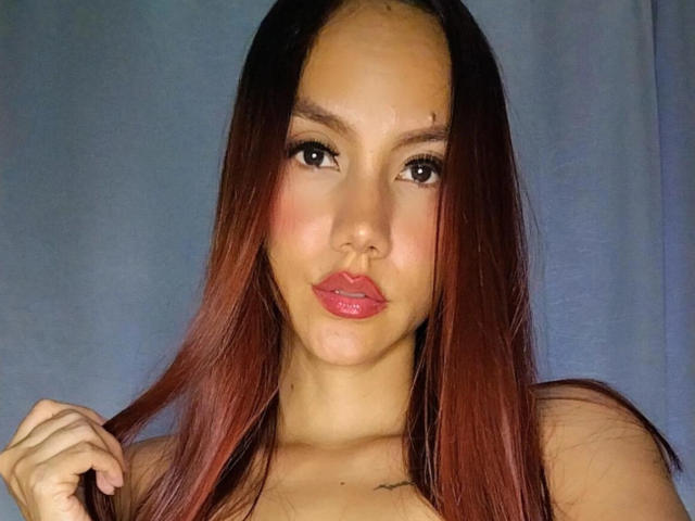 NormaEttie with long red-brown hair, pink lips, and light skin, holding her hair with a slight pout, showing small tits against a blue background.