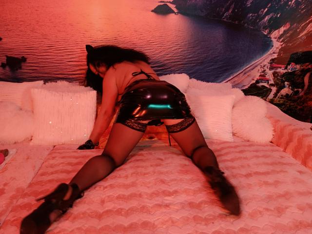 AmberSunshine on all fours, wearing black latex booty shorts, lace garters, and high heels, with her ass up and back arched, in a red-lit room.