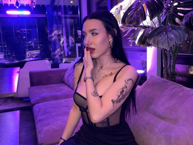 NikkiGoldX with long black hair, red lipstick, and tattoos, sitting on a purple sofa, touching her face, wearing a black sheer dress, exposing her small tits.