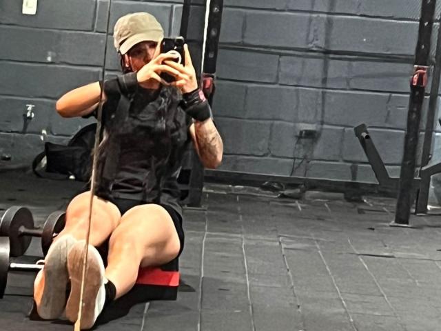 SteffanyTaylor with a cap, black shirt, and shorts, sitting on a bench, holding a phone, bare feet up, gym, weight rack, and dumbbells in the background.
