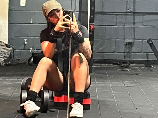 SteffanyTaylor is sitting on a weight bench, taking a mirror selfie, wearing a baseball cap, black shirt, black shorts, white sneakers, and wristbands, in a gym.