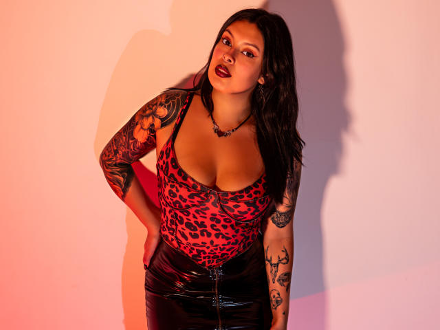 GabyJoy with large tits, wearing a red leopard print top and black shiny latex skirt, has tattooed arms and a confident pose, standing against a softly lit wall.