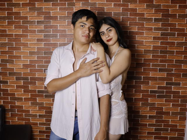 SofiNdLogan with dark hair, in white lace dress, stands behind a chubby man with black hair, wearing a pink shirt, hand on his chest, brown brick wall in background.
