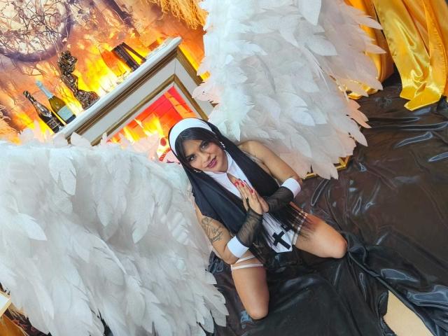 Mia Bellucci, avec des ailes d'ange, portant une tenue de nonne, des gants noirs et un chapeau blanc, agenouillée sur un tissu noir, montrant ses seins et un tatouage sur son bras gauche..