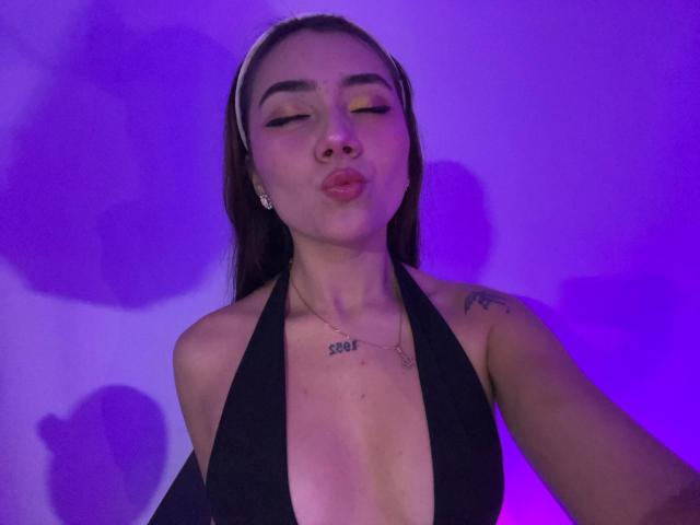 AlannaJonson with pouty lips, wearing a black halter top, showing her tits, tattoo on chest, brown hair, white headband, purple light, eyes closed, and small earrings.