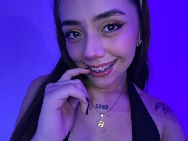 AlannaJonson with long brown hair, smiling, showing silver braces, black halter top, gold crown necklace, tattoo on right shoulder, purple-blue lighting, finger near mouth.