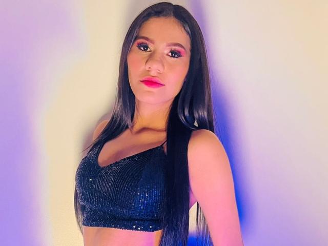 AmeliaSexx with long black hair, pink eyeshadow, and red lipstick, wearing a sparkly black crop top, showing her small tits and flat ass, against a blurred background.