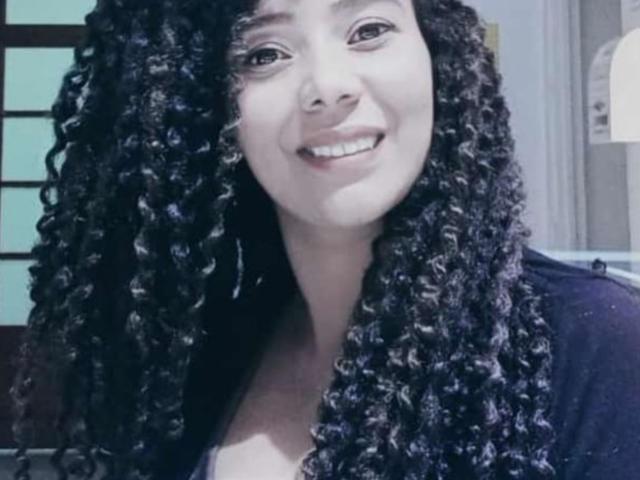 KenyaKarath with curly black hair, smiling, wearing a dark top, showing her white teeth, and a cute face, standing in a well-lit room.