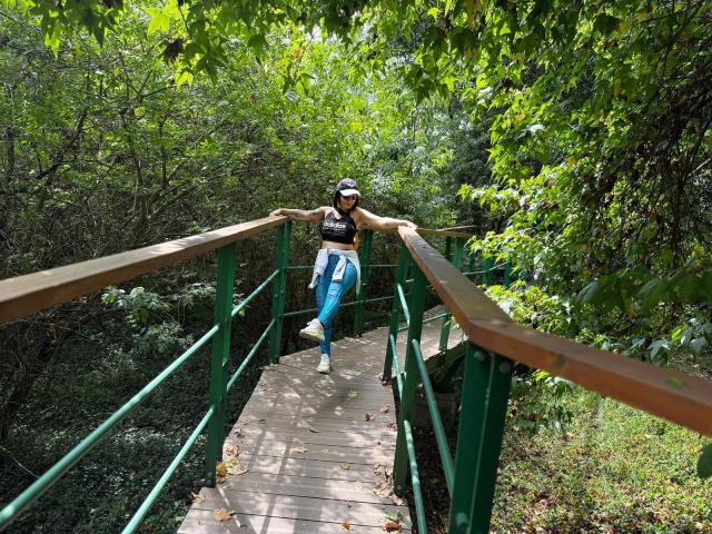 SaraleePoison leans on a green wooden bridge, wearing a black crop top, blue jeans, white sneakers, and a cap, with green leaves and trees in the background.