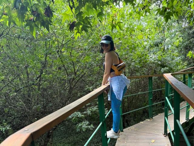 SaraleePoison with a black top, blue jeans, white sneakers, black visor, and guitar backpack, standing on a green wooden bridge, looking back over her shoulder.