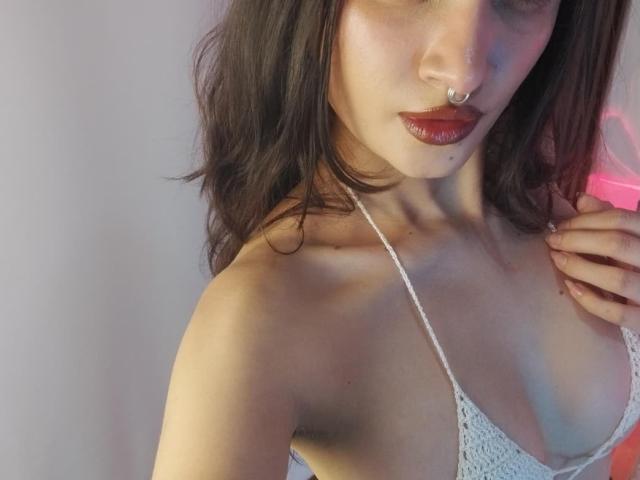 CandyyClair with wavy brown hair, red lipstick, nose ring, and white lace bralette showing her small tits. She's holding her hair, looking at the camera.