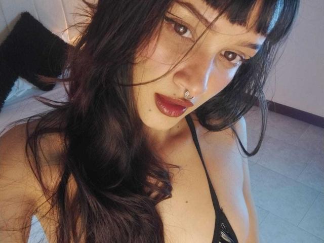 CandyyClair has long, dark, wavy hair, brown eyes, red lipstick, a nose ring, and is wearing a black strappy top, showing her small, perky tits.