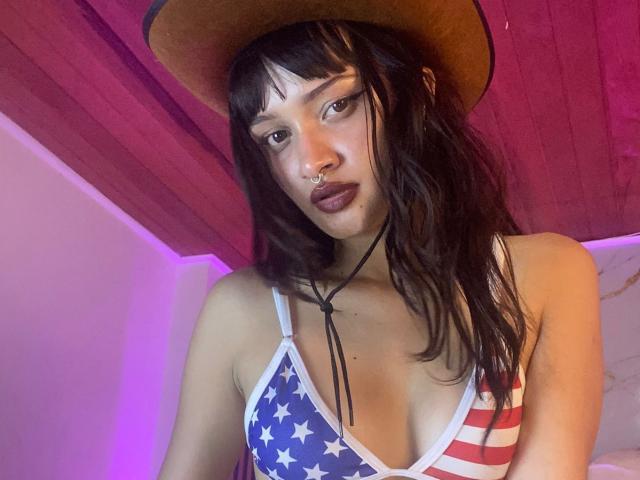 CandyyClair with wavy brown hair, wearing a cowboy hat, nose ring, and dark lipstick, in a star-spangled tank top, showing her tits, with purple lighting.