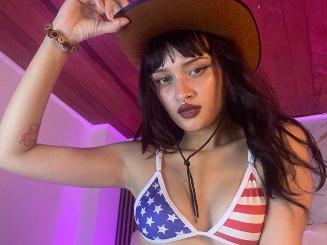 CandyyClair with dark hair, wearing a cowboy hat, star-spangled bikini top, nose ring, and black lipstick, holding the hat, showing small tits, and a tattoo on her arm.