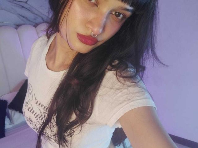 CandyyClair with long black hair, red lipstick, nose ring, and white t-shirt, showing a slight smile, her tits partially visible, in a purple-lit room.