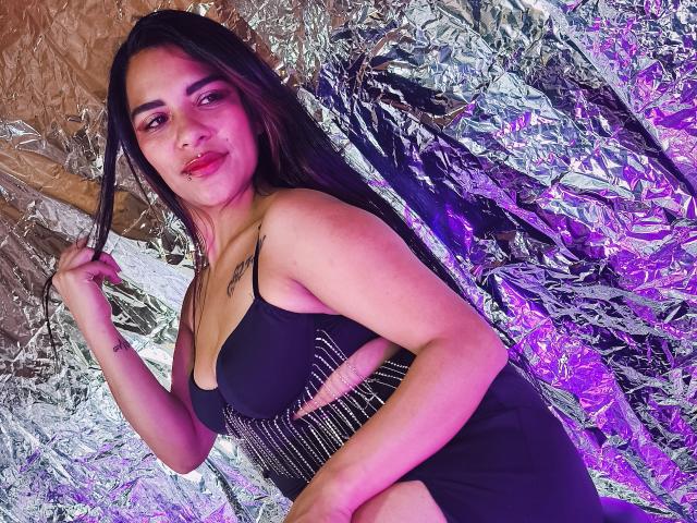 Mia Bellucci met lang zwart haar, roze oogmake-up en een zwarte jurk met metalen accenten, die een haarstreng vasthoudt, lachend, waarbij haar borsten en een tatoeage op haar pols zichtbaar zijn..