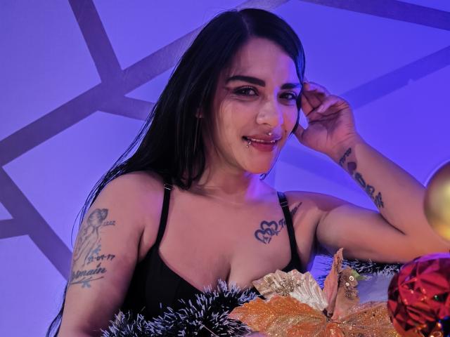 Mia Bellucci mit schwarzen Haaren, Tätowierungen an den Armen und einem Nasenring, trägt einen schwarzen BH, lächelt, hat eine Hand im Haar, auf dem Tisch befinden sich Brot und eine rote Dekoration, blaues Licht..