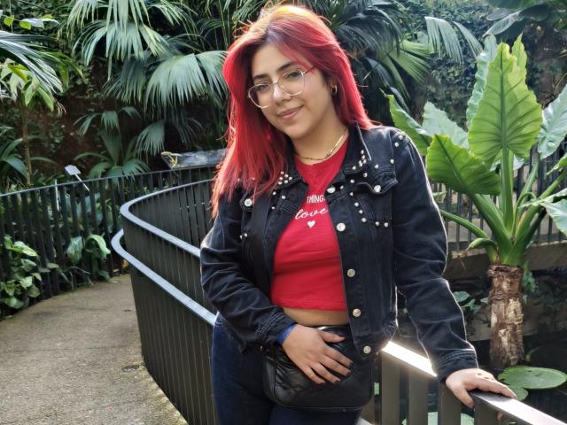 RousseColliinss with red hair, glasses, black studded jacket, red crop top, and blue jeans, holding a black purse, leaning on a black railing in a lush green setting.