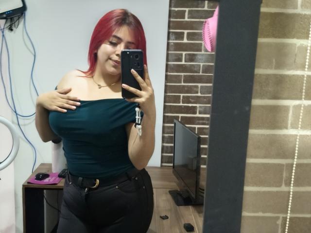 RousseColliinss con capelli rossi, top verde con una spalla scoperta, pantaloni neri, che tiene un telefono, sorridente, che copre i piccoli seni con la mano, selfie allo specchio, sfondo con un muro di mattoni, spazzola per capelli rosa nello specchio..