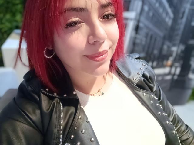 RousseColliinss with red hair, black leather jacket with studs, white top, hoop earrings, and a small necklace, smiling with pink lipstick. Boobs partially visible under jacket.