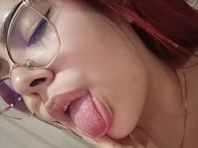 RousseColliinss with pink tongue out, blue eyeshadow, and glasses, sticking finger in mouth, red hair, fair skin, and slightly open lips. Her tongue is wet and she's looking up.