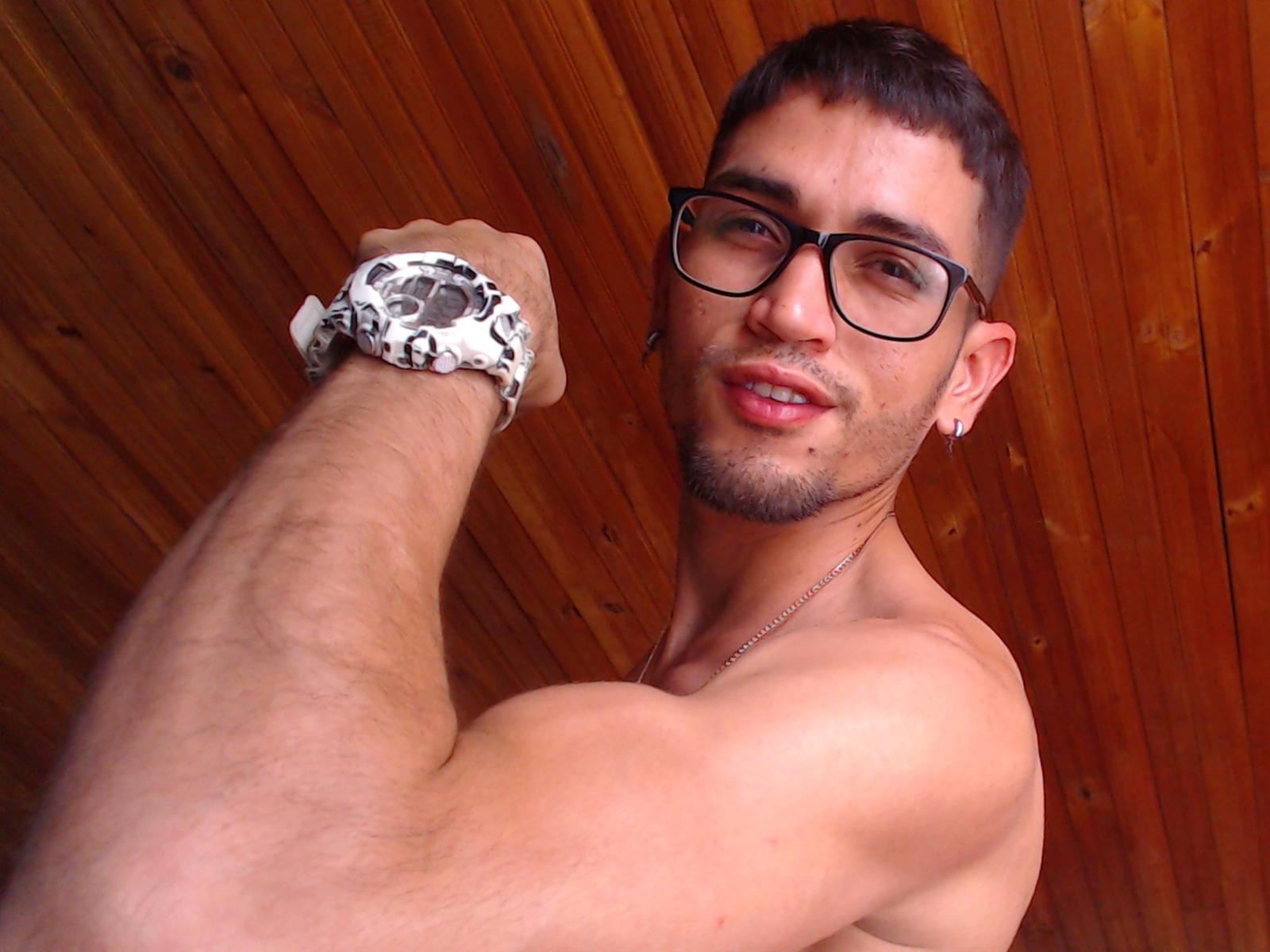 Akaza69, shirtless, with glasses, short brown hair, and a beard, flexing his muscular arm, wearing a white and black patterned wrist scrunch, smiling, wooden ceiling in background.