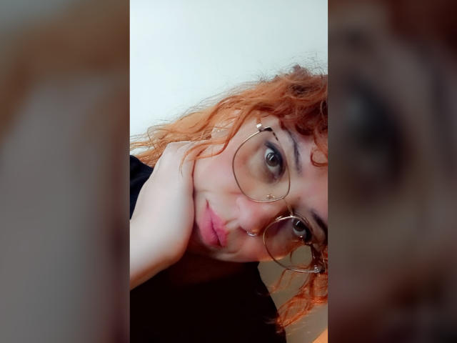 Close-up of RosseHot6.9 with curly, orange hair and round, wire-frame glasses. RosseHot6.9 has fair skin and is resting their head on their hand, looking directly at the camera with a slight, neutral expression. RosseHot6.9's eyebrows are thick and dark, and they have subtle pink lipstick. The background is blurred, focusing on RosseHot6.9's face and glasses. The image has a soft, natural light. RosseHot6.9's hair is slightly messy, adding a casual vibe.