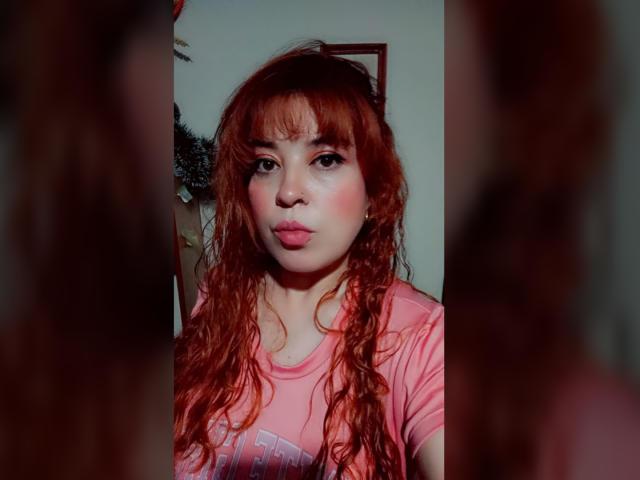 RosseHot69 has wavy, red hair and fair skin. She wears a pink t-shirt with a graphic. Her face is centered, slightly blurred in the foreground, with her reflection on the right. RosseHot69's lips are pouty, and she has dark, thick eyebrows. The lighting is even, with a subtle shadow on her left cheek. The image is a photograph with a shallow depth of field.