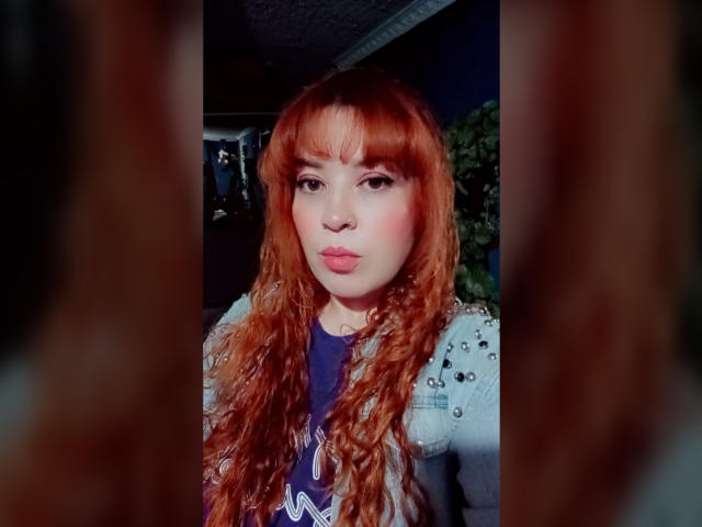 RosseHot69 has long, curly, red hair with bangs, and fair skin. She's pouting with pink lipstick. She wears a white, studded jacket over a blue shirt with white text. Her makeup includes dark eyeliner and pink blush. The image has a blurred brown border on both sides. The lighting highlights her face and hair, making them stand out. The background is dark and indistinct. The overall style is modern and slightly provocative. The image has a soft, almost digital filter effect.