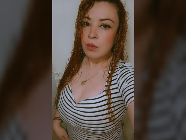 RosseHot69 ha i capelli castano ricci e mossi, indossa una maglietta a righe che mette in mostra il suo seno prosperoso, con una collana d'oro e un trucco leggero, guardando direttamente la fotocamera..