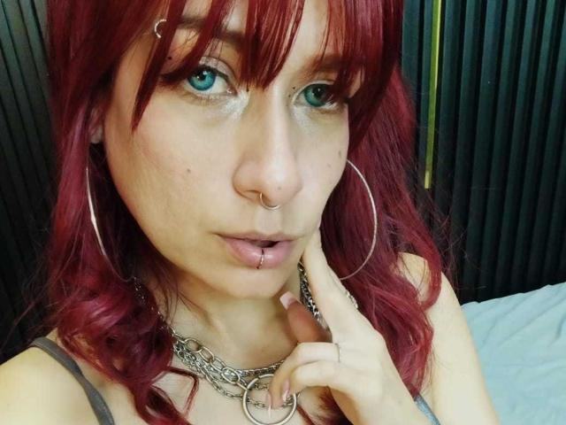LolyYunno with red hair, blue eyes, nose ring, lip piercing, and hoop earrings, wearing a chain necklace, touching her lower lip, looking at the camera.