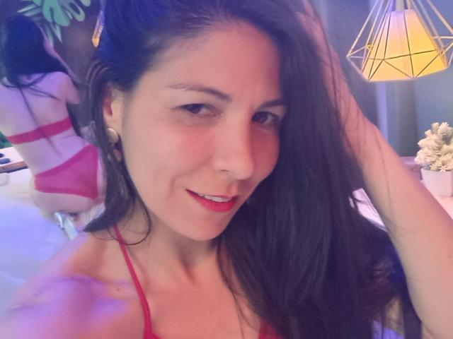 EliseBell a de longs cheveux foncés, du rouge à lèvres, et un léger sourire. Elle porte un haut rouge à bretelles fines, et a une main dans ses cheveux, mettant ainsi en valeur sa poitrine..